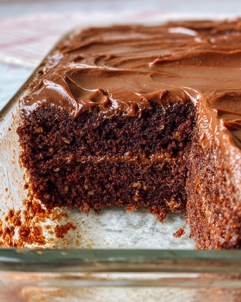 The image shows a close-up of a rich chocolate cake in a glass baking dish on a white marbled texture surface. The cake has two thick layers of moist, dark brown chocolate sponge with visible small nut pieces, and between and on top of these layers is a smooth, glossy layer of chocolate frosting. The frosting on top is shiny and evenly spread, with gentle swirls, and the cake looks very soft and gooey near the edges. The glass dish has some chocolate residue on the sides, adding to the homemade feel. photo taken with an iphone --ar 4:5 --v 7