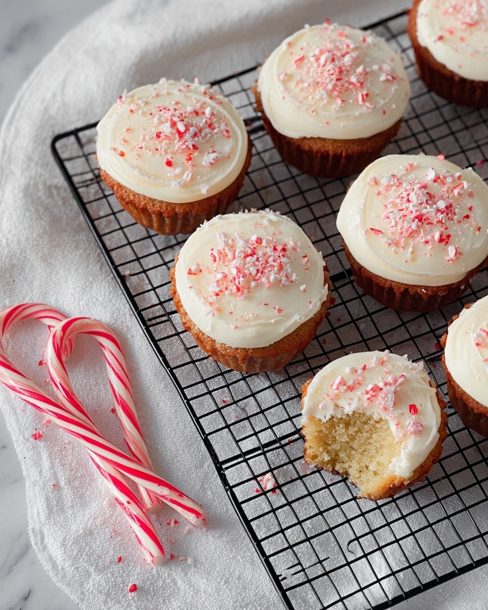A close-up of a cupcake with one layer of light brown cake in a silver wrapper at the bottom, topped with one thick, smooth layer of creamy white frosting spread evenly on top, sprinkled with small red and white crushed candy pieces. The cupcake is on a black cooling rack placed on a white marbled surface, with other similar cupcakes blurred in the background. Photo taken with an iphone --ar 4:5 --v 7