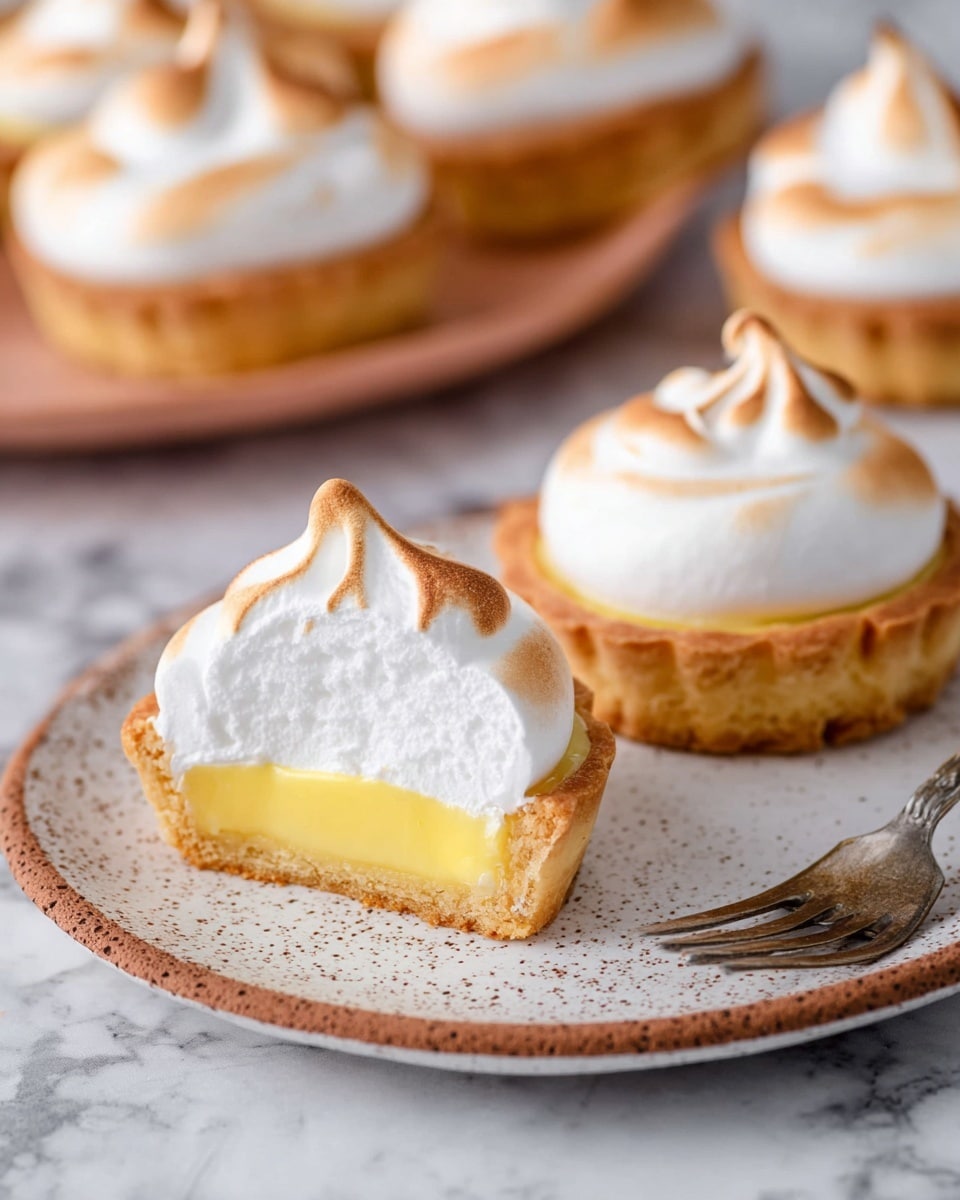 The image shows two mini lemon meringue tarts on a round white plate with a speckled brown pattern, placed on a white marbled surface. Each tart has a golden-brown crust filled with a smooth, bright yellow lemon custard layer, topped with a fluffy, white meringue layer that is lightly toasted to a soft golden brown at the peaks. One tart is cut in half, showing the clear separation of the three layers: the crust bottom, the creamy lemon filling middle, and the thick meringue top. In the background, more whole tarts are arranged on the white marbled surface, blurred softly to keep focus on the tarts on the plate. A vintage silver fork is placed next to the plate. photo taken with an iphone --ar 4:5 --v 7