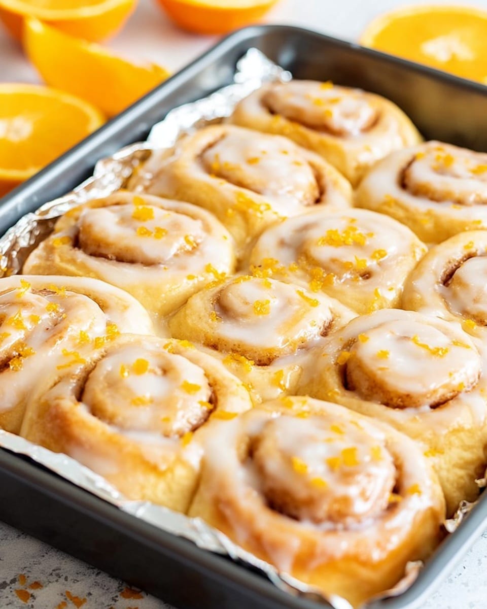 A close-up view of a baking pan filled with nine orange-glazed cinnamon rolls arranged in three rows and three columns. Each cinnamon roll shows soft layers of dough rolled tightly, with a light orange glaze evenly spread on top, giving a shiny and sticky texture. Orange zest bits are scattered over the glaze, adding small bright orange specks on the creamy surface. The baking pan is lined with shiny foil, with the rolls sitting close to each other, displaying light golden-brown dough layers beneath the glaze. The scene has blurred orange slices in the background and is set on a white marbled texture. Photo taken with an iphone --ar 4:5 --v 7