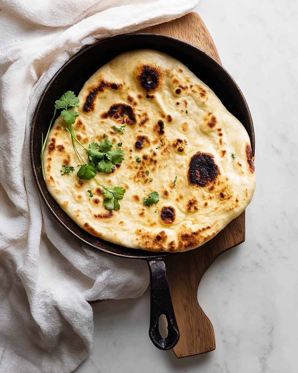A single round piece of naan bread is shown in a dark pan with a handle, resting on a light wooden board on a white marbled surface. The naan has a golden-brown color with several darker char marks scattered unevenly across its slightly puffed surface. It appears soft and slightly puffy with a bubbly texture. On top, there are a few sprigs of fresh green cilantro placed for garnish. A white cloth is partly visible on the left side of the board. photo taken with an iphone --ar 4:5 --v 7