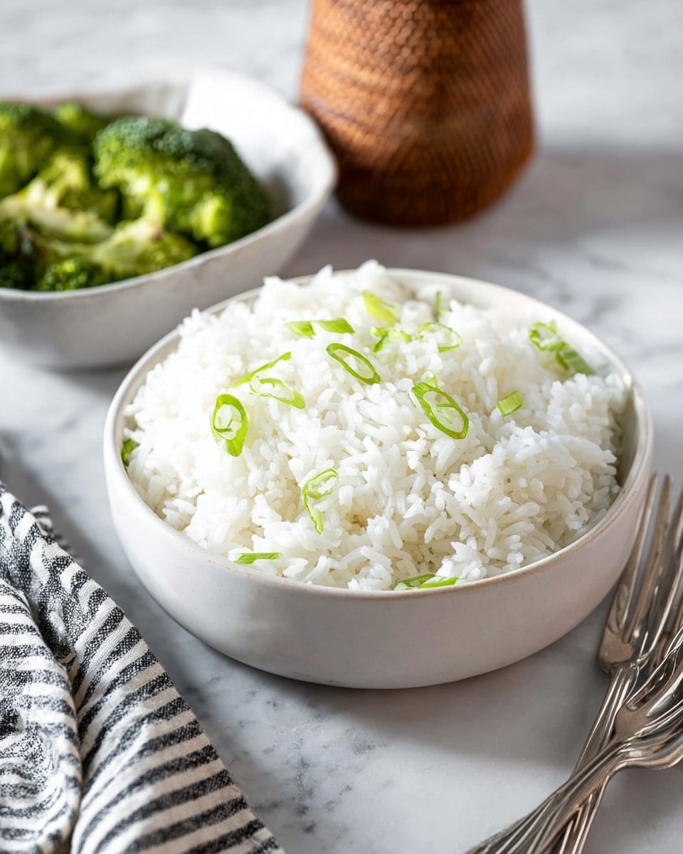 A white bowl filled with a single layer of fluffy white rice topped with scattered thin green slices of scallion, placed on a white marbled surface next to a white bowl of green broccoli florets in the background, with an out-of-focus brown woven bottle and black and white striped cloth nearby, two silver forks resting beside the bowls photo taken with an iphone --ar 4:5 --v 7