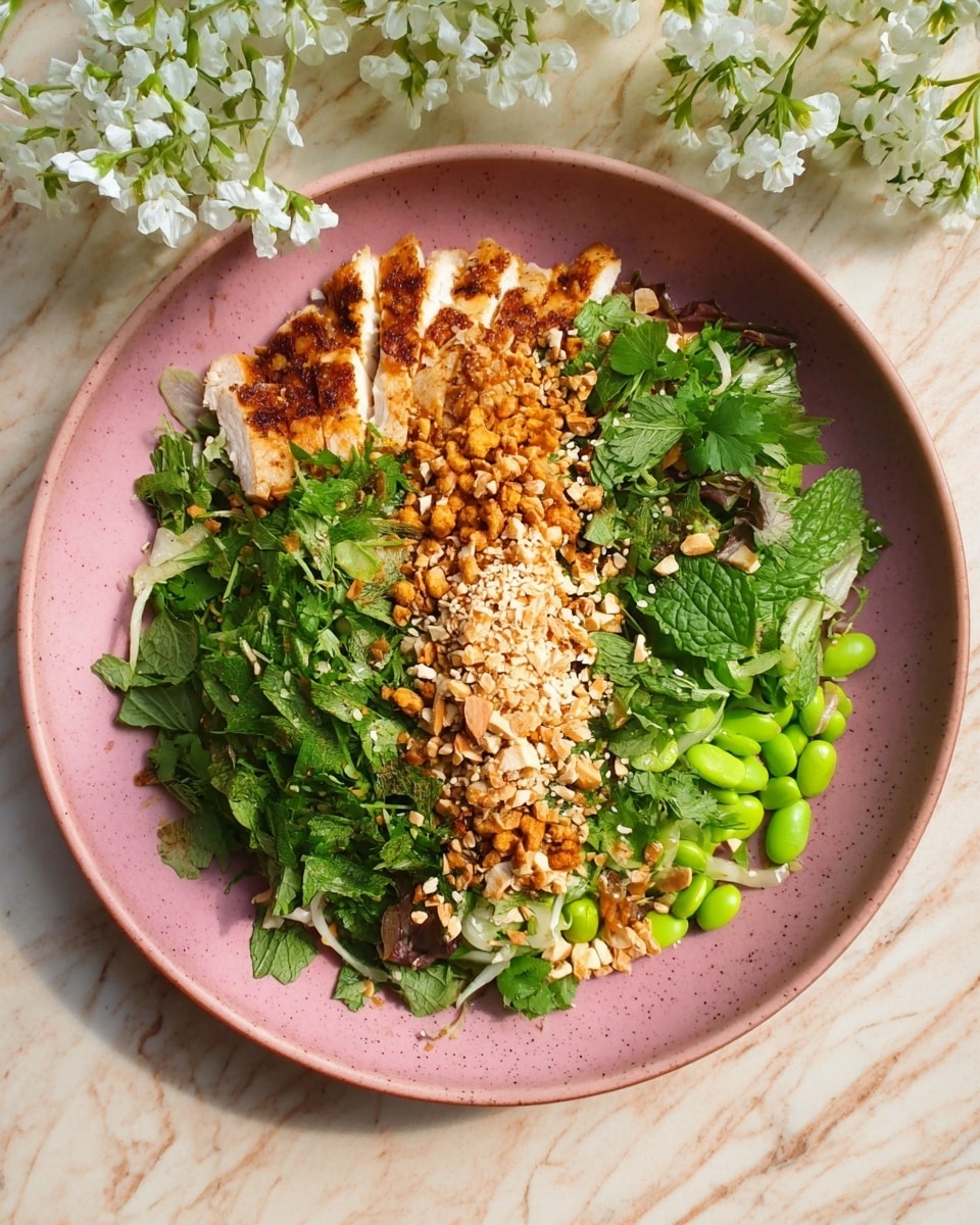 A pink ceramic plate holds a colorful dish with multiple layers. Starting from the left, there is a layer of sliced grilled chicken with a golden-brown crust. Next to it, fresh green herbs dyed in various shades cover most of the plate, including shredded mint, cilantro, and other leafy greens. In the center, a strip of chopped roasted peanuts and white sesame seeds runs vertically, adding a crunchy texture and light tan colors. Some bright green edamame beans and sliced fresh herbs add more vibrancy to the right side. The plate is placed on a white marbled textured surface with white flowers visible at the top corner. Photo taken with an iphone --ar 4:5 --v 7