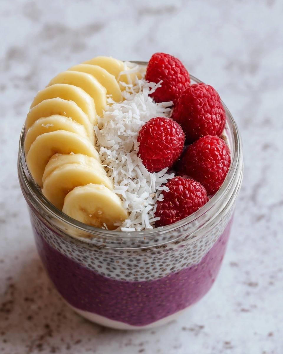 This image shows a two-layered chia pudding in a clear glass jar placed on a white marbled surface. The bottom layer is purple with visible chia seeds, smooth and evenly spread. The top layer has a curved row of creamy yellow banana slices on one side, a few white shredded coconut flakes scattered beside them, and four bright red raspberries lined up next to the banana slices on the other side. The colors contrast nicely, making the dish look fresh and colorful. Photo taken with an iphone --ar 4:5 --v 7