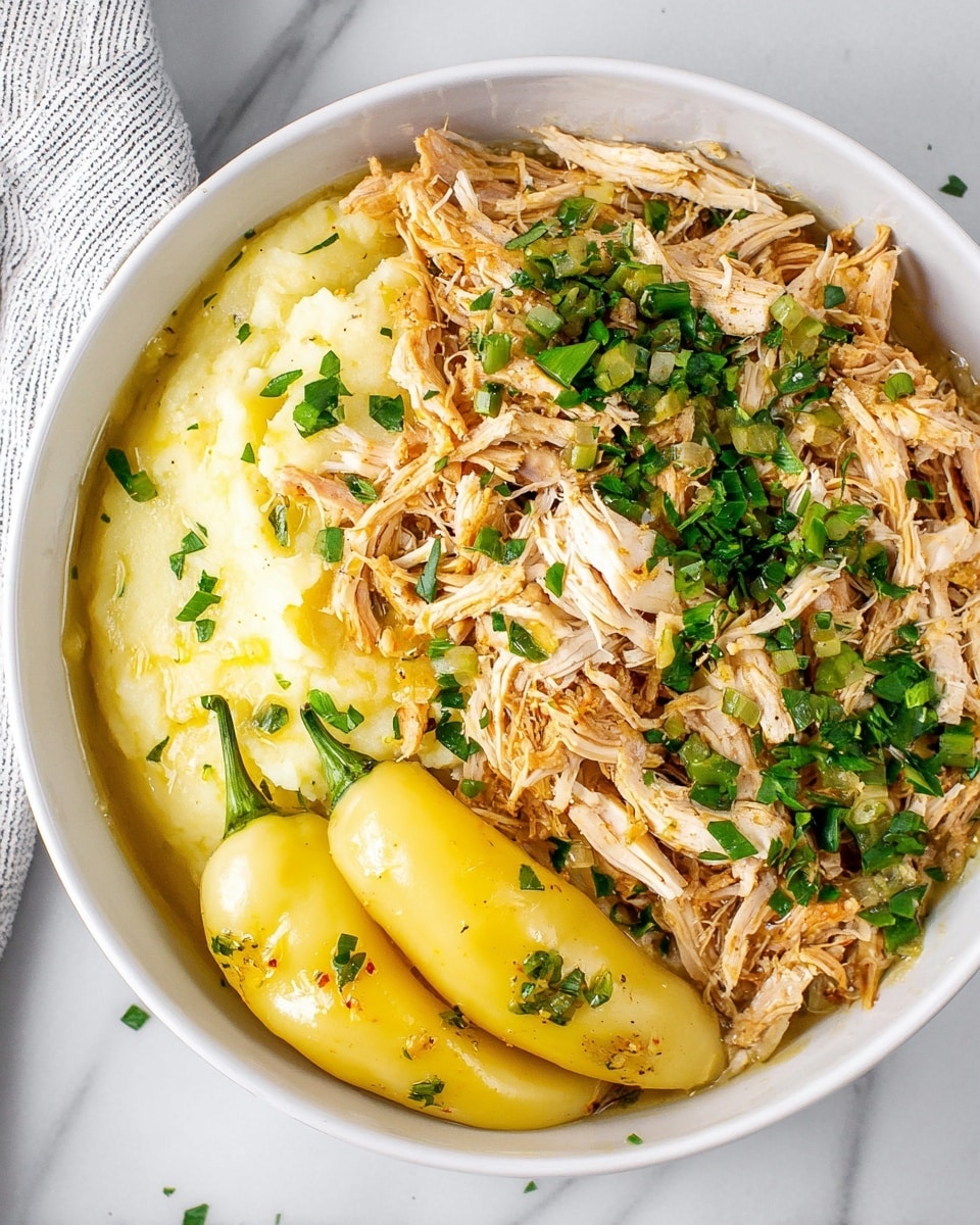 A white bowl contains three main layers: on the left and center, shredded light brown chicken mixed with green chopped herbs, scattered all over the meat; on the right, a smooth and creamy yellow layer of mashed potatoes; at the bottom, two pale yellow whole peppers resting side by side, each with a shiny surface and sprinkled with some herbs. The bowl is set on a white marbled surface with a striped cloth partially visible at the top left corner. photo taken with an iphone --ar 4:5 --v 7