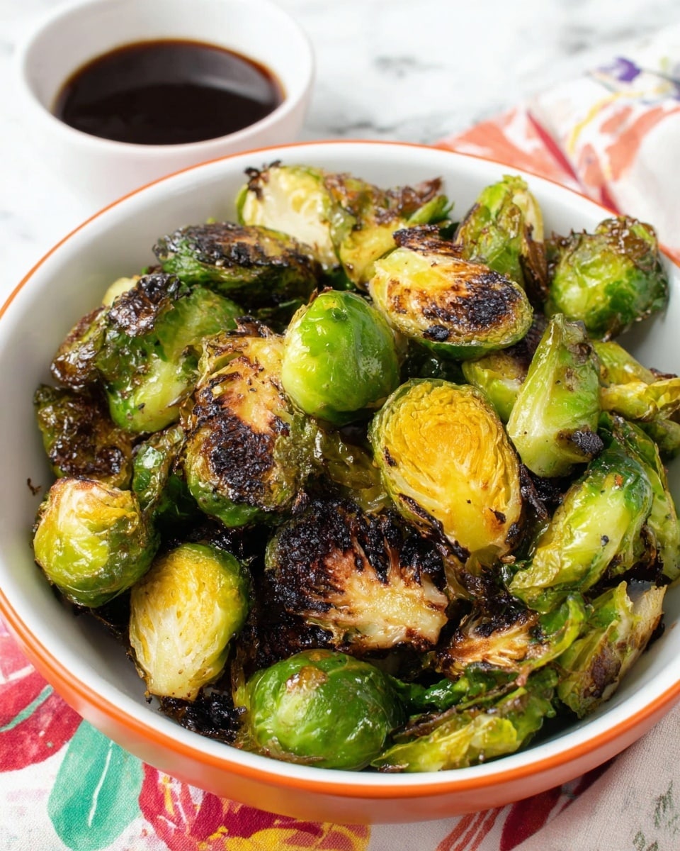 A white bowl with an orange rim is filled with roasted Brussels sprouts, showing several layers of green outer leaves that are charred dark brown and crispy, mixed with bright green and yellow soft inner parts of the sprouts, some cut in half revealing their layered texture. To the side, there is a white bowl containing dark brown dipping sauce, all placed on a white marbled surface with a colorful, partially visible cloth underneath. photo taken with an iphone --ar 4:5 --v 7