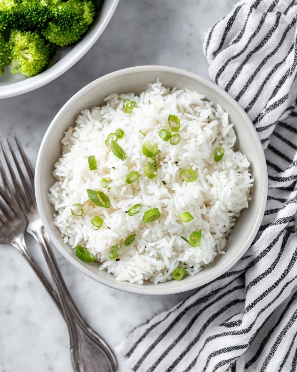 A white bowl filled with fluffy white rice, topped with small pieces of bright green sliced scallions scattered evenly across the surface. The texture of the rice looks soft and slightly sticky, with the scallions adding a fresh, crisp contrast. The bowl sits on a white marbled surface with a striped black and white cloth napkin on the right side and two silver forks placed casually to the left. In the background, partially visible, there is another white bowl containing green broccoli florets. photo taken with an iphone --ar 4:5 --v 7