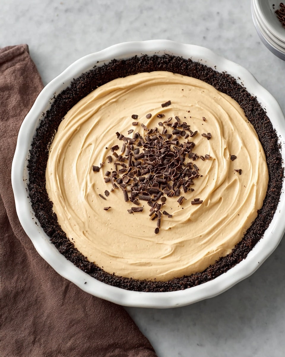 A round pie with three visible layers sits on a white marbled surface: the bottom layer is a dark chocolate crumb crust with a rough texture, forming a thick edge around the pie; the middle layer is a smooth, creamy light tan filling that is spread evenly but with soft swirls on top; the center is decorated with small dark chocolate curls scattered neatly in a pile. The pie is placed in a white fluted ceramic dish, and a brown cloth is partially visible in the corner. Photo taken with an iphone --ar 4:5 --v 7
