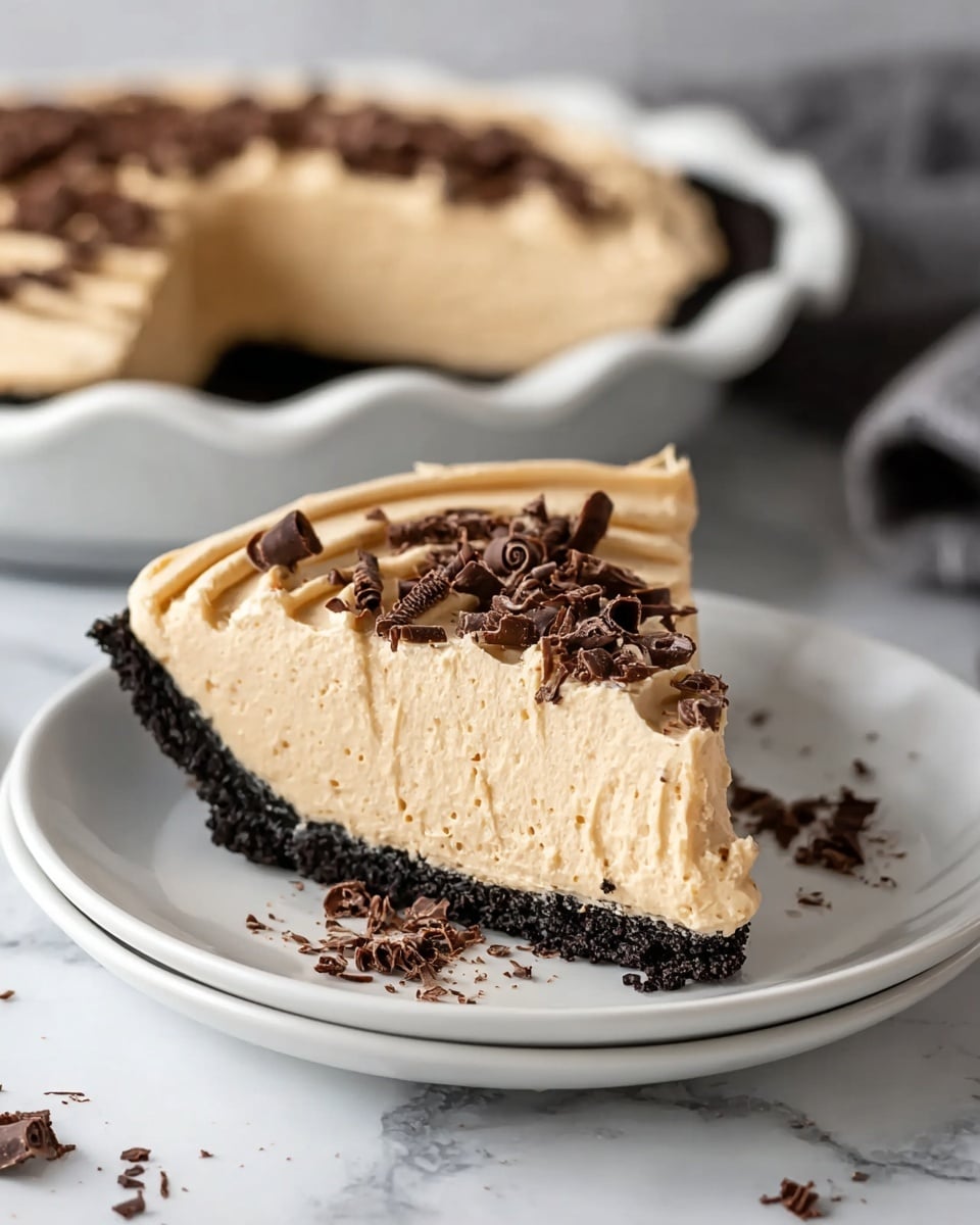 A slice of pie sits on a white plate with a dark crumbly crust forming the bottom layer, topped by a thick, light tan creamy filling as the second layer. The top layer is smooth with slight swirls and decorated with scattered dark chocolate curls. In the background, the rest of the pie with the same layers and chocolate curls is visible in a white pie dish, placed on a white marbled surface. There are small crumbs and chocolate shavings scattered around the plate and surface. photo taken with an iphone --ar 4:5 --v 7