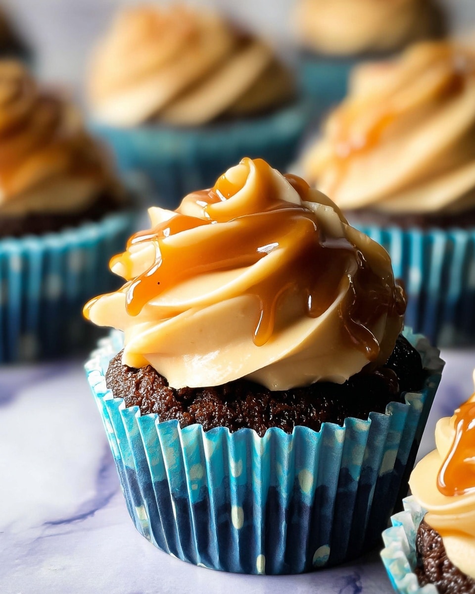 A close-up of a dark chocolate cupcake in a blue patterned paper liner sitting on a white marbled surface. The cupcake has one thick layer of smooth, light caramel-colored frosting that is piped in a swirled spiral shape with a slight peak in the middle. Drizzled on top is a glossy, darker caramel sauce adding a shiny texture and rich amber color to the creamy frosting. Other similar cupcakes sit blurred in the background. Photo taken with an iphone --ar 4:5 --v 7