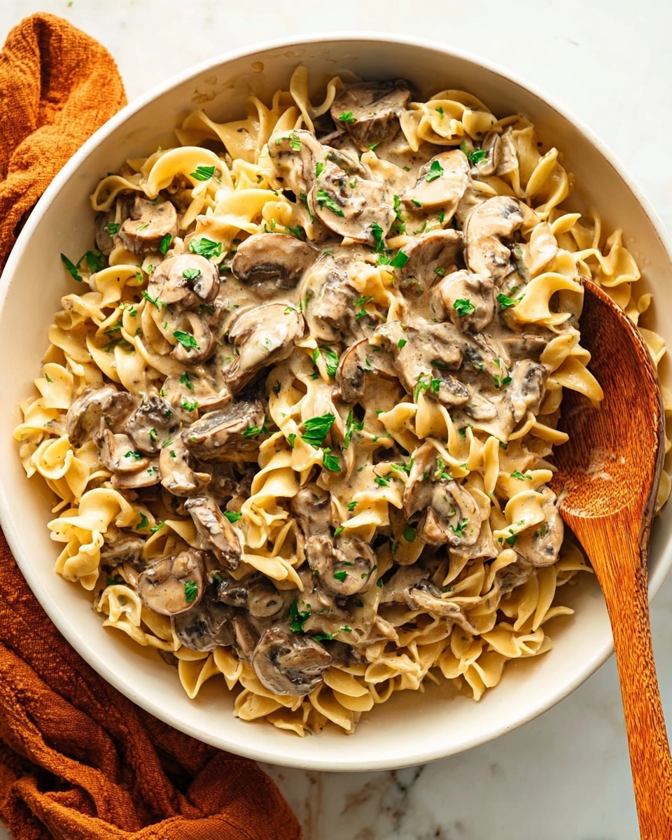 A white bowl filled with soft, curly egg noodles layered with creamy mushroom sauce mixed with sliced, cooked brown mushrooms scattered throughout. The sauce is smooth with a light beige color, coating the noodles evenly. Small green parsley pieces are sprinkled on top, adding a fresh contrast to the warm tones of the dish. A wooden spoon is partially buried in the noodles on the right side of the bowl. The bowl rests on a white marbled surface with a folded orange cloth nearby. photo taken with an iphone --ar 4:5 --v 7