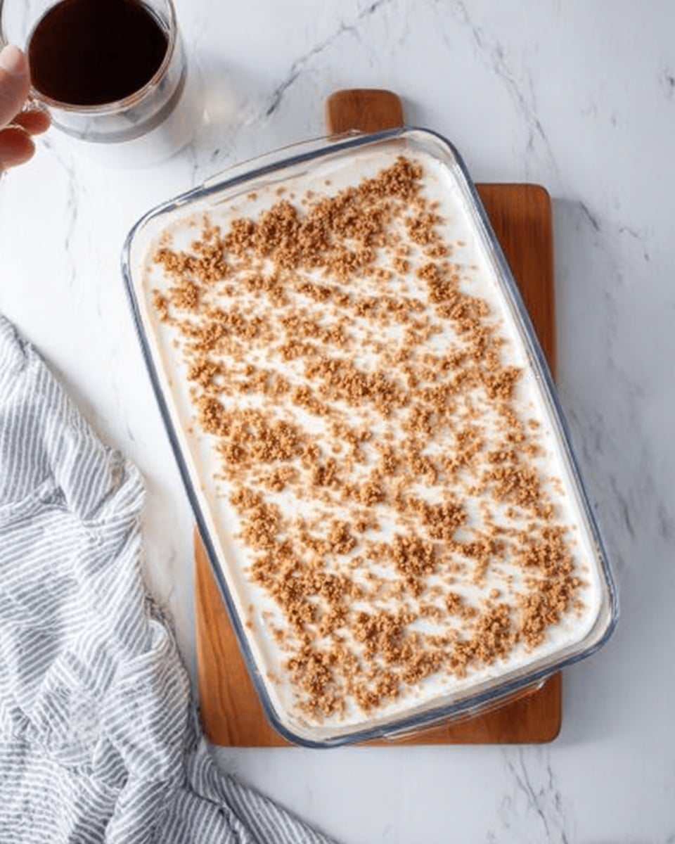A clear glass rectangular dish filled with a creamy white dessert topped evenly with small brown crumbs, placed on a wooden board over a white marbled surface. The dessert looks smooth with a soft texture and a single layer of crumb topping scattered across the top. A woman's hand holding a clear glass cup is partially visible at the top left, and part of a white and gray striped cloth is on the bottom left. Photo taken with an iphone --ar 4:5 --v 7