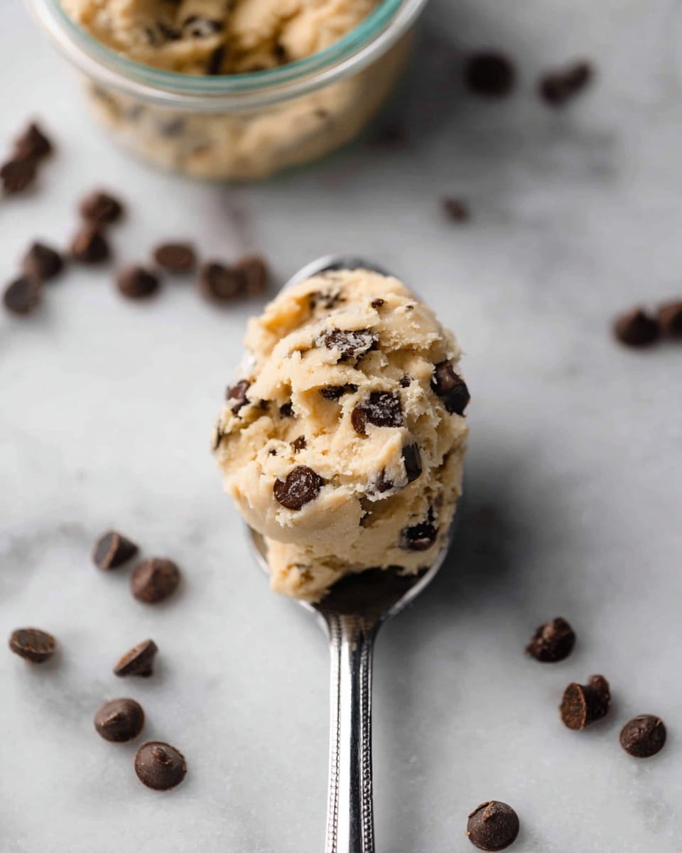 A close-up view of a spoon holding a scoop of light beige cookie dough mixed with dark brown chocolate chips, resting on a white marbled surface; in the background, a small clear container filled with more cookie dough and scattered chocolate chips around it. Photo taken with an iphone --ar 4:5 --v 7