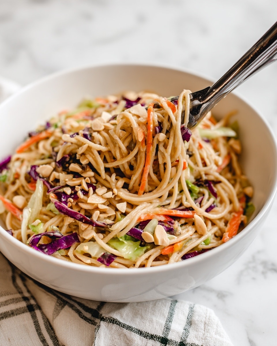 The image shows a white bowl filled with a colorful noodle salad. The salad has multiple layers with light tan cooked noodles as the base, mixed with thin strips of green cabbage, orange carrot slices, and small pieces of purple cabbage. There are also almond slices and some sunflower seeds sprinkled throughout, adding texture. A pair of black and silver tongs is resting inside the bowl, lifting a portion of the salad, with a white and green checked cloth underneath the bowl. The background surface has a white marbled texture. photo taken with an iphone --ar 4:5 --v 7