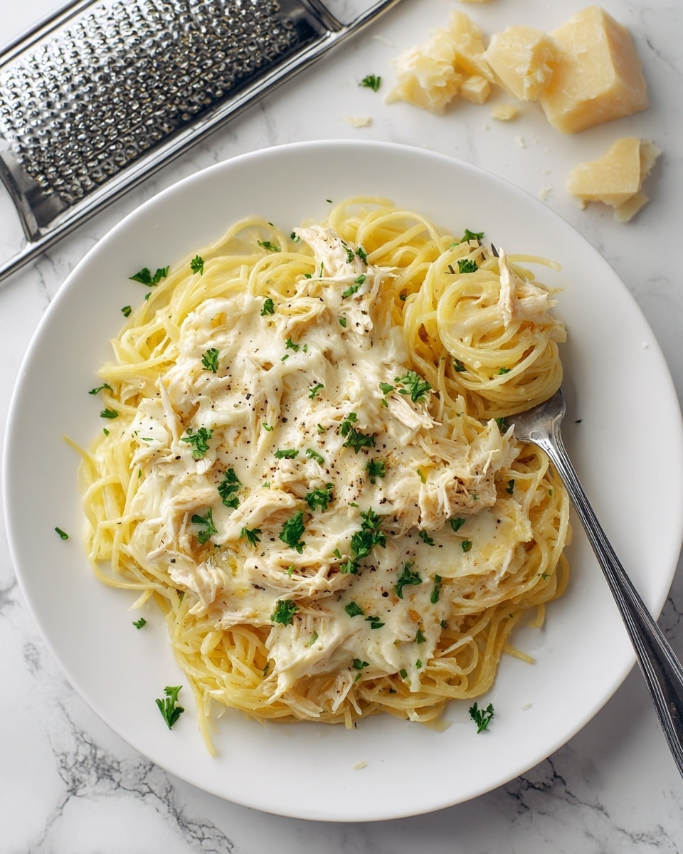 A white plate filled with thin, yellow spaghetti pasta arranged in loose nests, topped with creamy, white Alfredo sauce mixed with shredded chicken pieces. The sauce is thick and slightly speckled with black pepper, while finely chopped green parsley is scattered over the top for color. A silver fork twirls a bite of spaghetti on the right side of the plate. In the background, there is a white grater with some chunks of cheese on top, all set on a white marbled texture. photo taken with an iphone --ar 4:5 --v 7