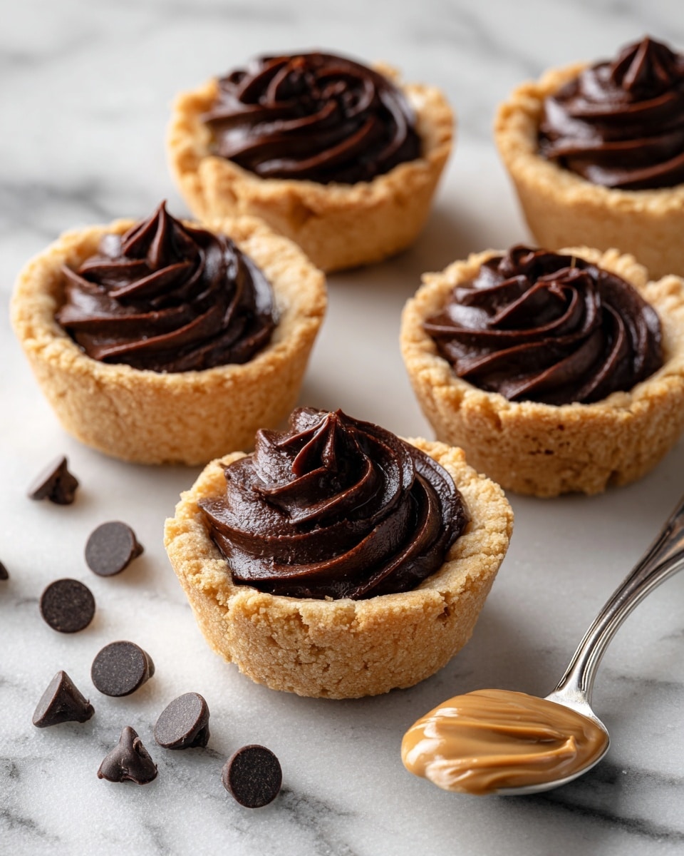The image shows five small cookie cups arranged on a white marbled surface, each filled with a thick, smooth layer of dark chocolate frosting swirled on top, creating a soft peak in the center. The cookie cups are golden brown with a slightly crumbly texture, forming a sturdy base and rim around the chocolate. Scattered around the cookie cups are several dark chocolate chips, and a silver spoon with a swirl of creamy peanut butter rests to the side, adding contrast in color and texture. The scene is softly lit, highlighting the glossy chocolate and the crumbly cookie details, photo taken with an iphone --ar 4:5 --v 7