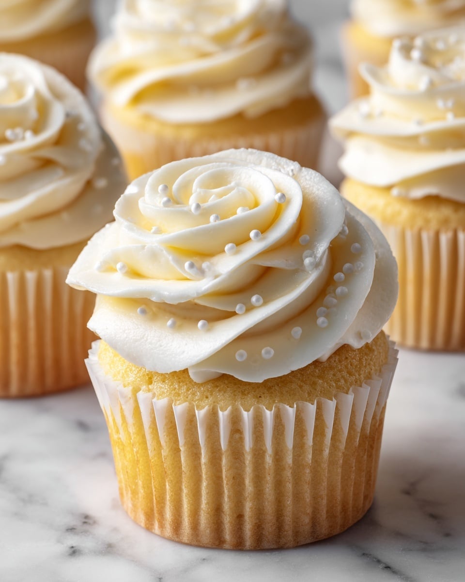 A close-up view of a vanilla cupcake with a light brown base in a white paper liner. On top, there is a thick swirl of creamy white frosting, decorated with small white sugar pearls. The cupcake is centered among several similar cupcakes in the background, all on a white marbled surface with a soft blue background. The frosting swirls have a smooth and fluffy texture, creating a soft, inviting look. photo taken with an iphone --ar 4:5 --v 7