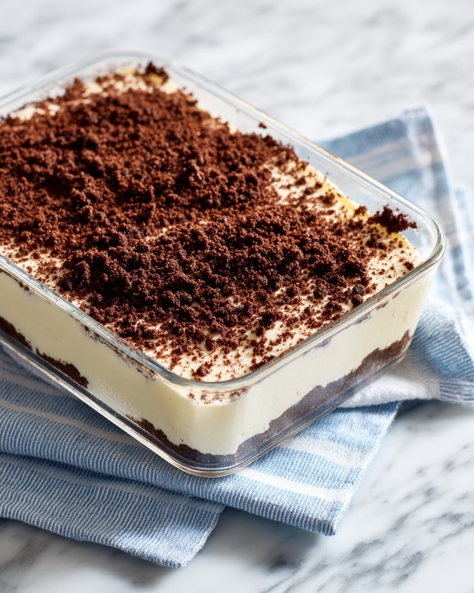 A clear glass rectangular dish filled with a dessert made of two main layers: the bottom layer is thick, creamy white, smooth and even, while the top layer is a generous, uneven scattering of dark brown, crumbly cookie crumbs covering the entire surface. The dish is placed on a white marble surface with a light blue striped cloth partially visible under it. photo taken with an iphone --ar 4:5 --v 7