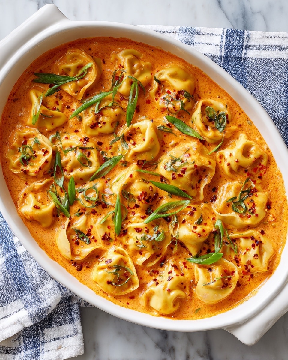 The dish shows a white rectangular baking dish filled with folded yellowish pasta sheets layered closely together, each piece soft and slightly curled at the edges. The pasta is covered in a thick, rich orange sauce with a creamy texture, pooling around the edges and between the pieces. Scattered on top are small bits of bright red chili flakes and cleaned slices of green onion rings, adding color contrast. There are also fresh green herbs sprinkled evenly across the surface. The overall look is warm and inviting with a mix of smooth sauce and textured toppings, sitting on a white marbled surface. photo taken with an iphone --ar 4:5 --v 7