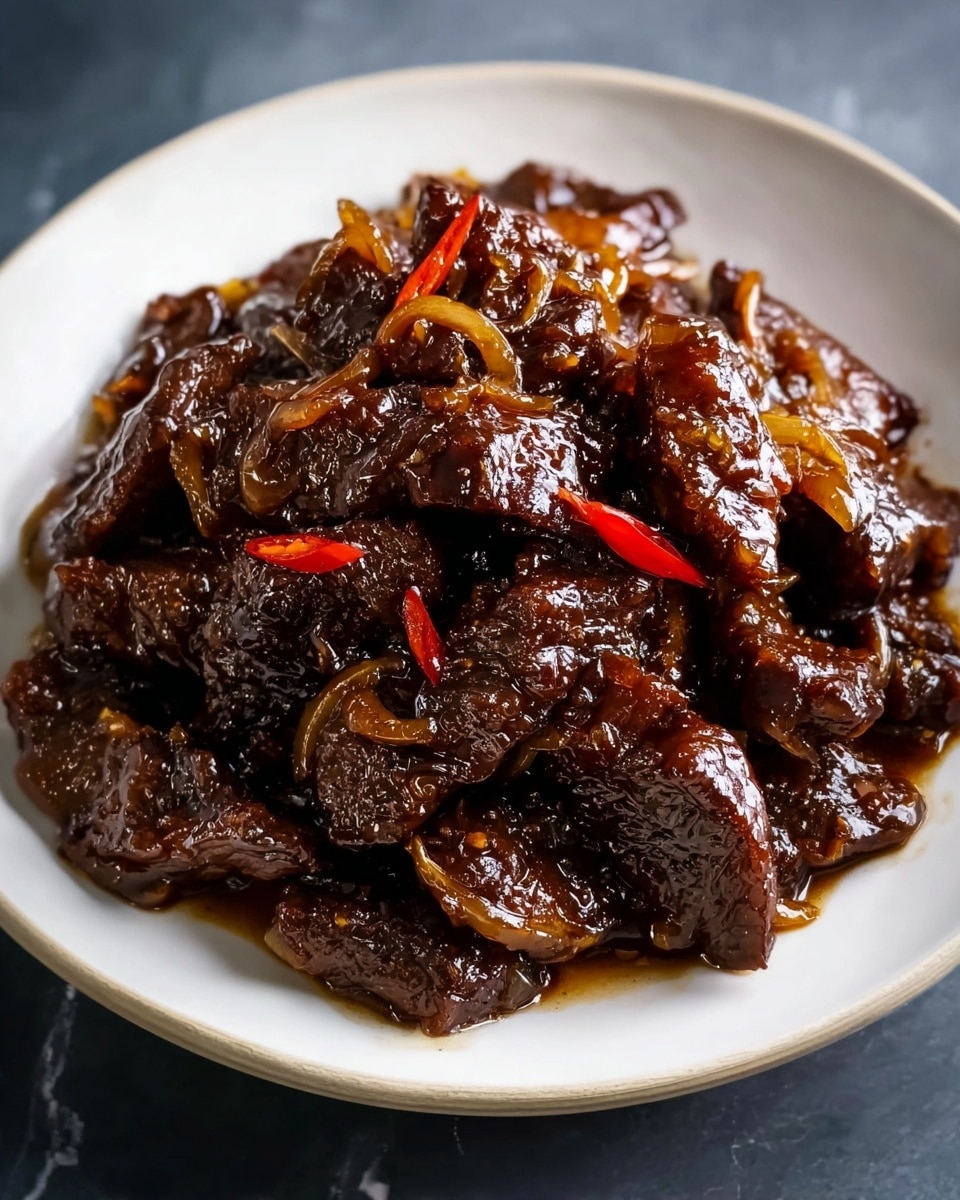A white plate filled with a pile of glossy dark brown beef pieces cooked in a sticky sauce, with thin caramelized onion slices mixed in and a few small red chili slices scattered on top. The beef looks tender and shiny, with a rich, thick coating of sauce that glistens under the light. The plate is placed on a white marbled surface. photo taken with an iphone --ar 4:5 --v 7