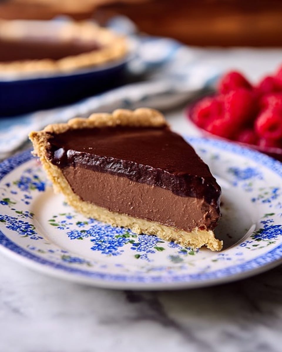 A slice of chocolate pie is shown with three layers: a light golden brown crust at the bottom, a thick, smooth dark brown chocolate filling in the middle, and a darker glossy chocolate layer on top. The slice rests on a round white plate with a blue floral pattern featuring clusters of small blue flowers and green leaves. In the background, there is a container of bright red raspberries blurred softly. The whole scene is set on a white marbled texture surface. photo taken with an iphone --ar 4:5 --v 7