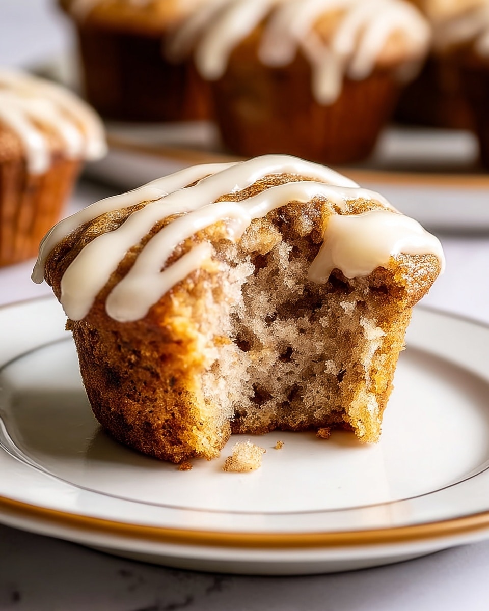 A close-up view of a single muffin with one bite taken out, showing its soft, moist, and slightly crumbly light beige inside with darker cinnamon specks throughout. The muffin has a golden brown crust that is textured and slightly rough around the edges. On top, there is a thick drizzle of smooth, creamy white icing creating a striped pattern. The muffin sits on a white plate with a thin gold rim, placed on a white marbled surface. In the blurred background, there are more muffins visible. photo taken with an iphone --ar 4:5 --v 7