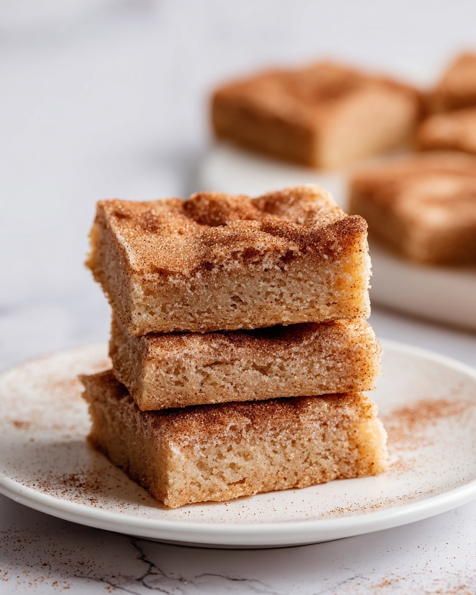 A stack of three square cinnamon sugar bars is placed on a white plate, sitting on a white marbled surface. Each bar has a light brown color with a soft, crumbly texture. The top layer of the bars is dusted generously with a mix of cinnamon and sugar that creates a speckled look of darker and lighter browns. The edges are slightly firm, while the inside appears moist with small air pockets. In the background, blurred squares of the same bars sit on the white marbled surface, enhancing the focus on the stacked bars. Photo taken with an iphone --ar 4:5 --v 7