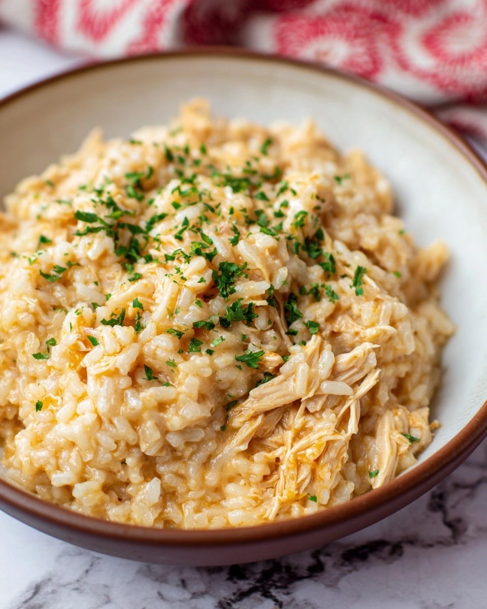 A close-up view of a creamy rice dish served in a white bowl with a brown rim, showing a single layer of soft, slightly sticky rice mixed with shredded chicken in a light orange sauce. The dish has finely chopped green herbs sprinkled evenly on top, adding small bright green accents. The texture looks smooth and moist, with the rice and chicken well combined. The bowl sits on a white marbled surface with part of a red and white patterned cloth visible in the blurred background. photo taken with an iphone --ar 4:5 --v 7