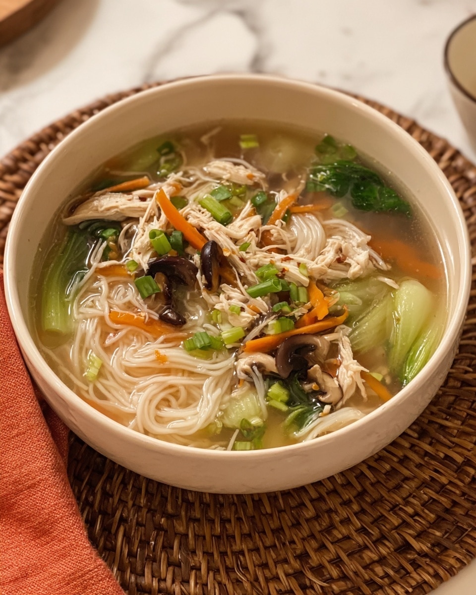 A white bowl filled with a clear broth that holds soft, pale noodles layered at the bottom, topped with a mix of light green bok choy leaves, thin orange carrot slices, and small dark brown mushroom pieces. Scattered on top are small pieces of shredded white chicken and chopped bright green scallions, all floating gently in the soup. The bowl is placed on a woven mat, with a white marbled surface in the background. photo taken with an iphone --ar 4:5 --v 7