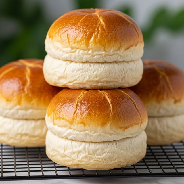 The image shows three soft, round bread rolls with shiny golden-brown tops and light, fluffy bottoms resting on white parchment paper. Each roll has a slightly puffy, layered texture with small peaks and valleys that create a cloud-like appearance. The bread's color gradually shifts from a deep amber on top to a pale cream at the base. In the blurred background, the greenery outside gives a fresh, outdoorsy feel. The surface beneath the parchment paper is a white marbled texture. photo taken with an iphone --ar 4:5 --v 7