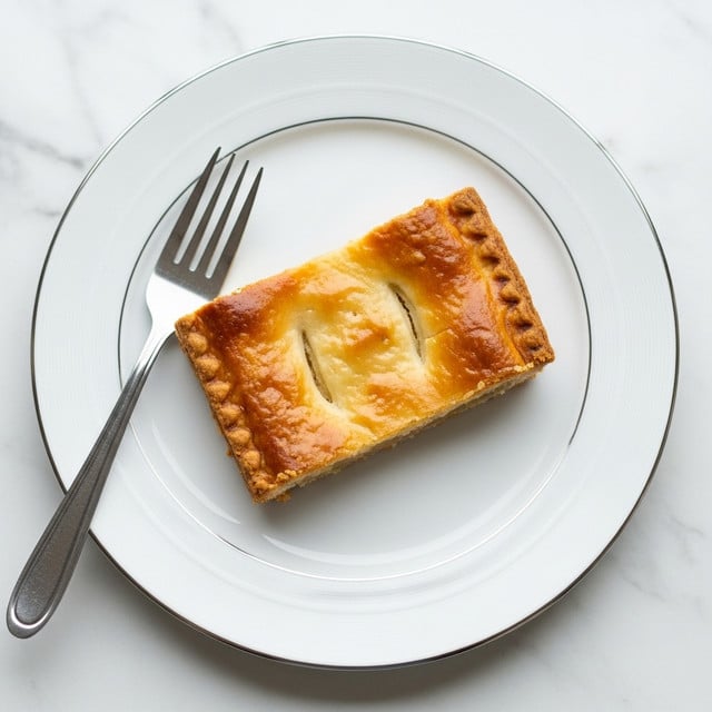 A single rectangular piece of golden brown pie sits in the center of a white plate with a thin silver rim. The pie has a smooth, slightly shiny top crust with small wrinkles and a light flaky texture visible. A silver fork rests on the left side of the plate, its prongs pointing toward the pie. The plate is placed on a white marbled surface, creating a clean and simple background. photo taken with an iphone --ar 4:5 --v 7
