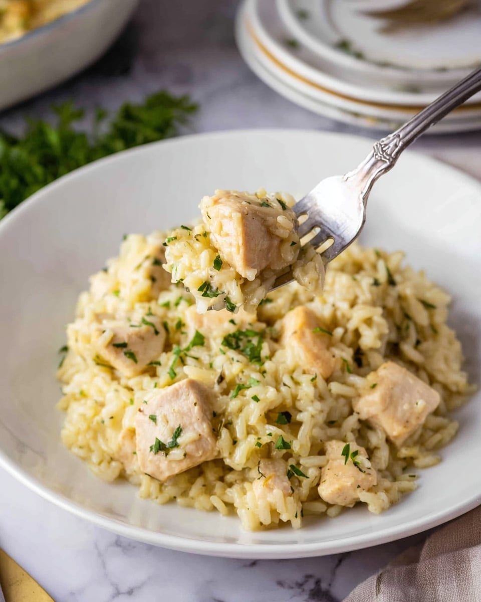 A white plate holds a serving of creamy rice mixed with tender chicken pieces. The dish has a soft texture with the rice cooked to a light golden color and the chicken cubes showing a pale, juicy look. Small bits of green herbs are scattered on top and mixed in, adding a fresh touch. A silver fork lifts a bite of chicken and rice above the plate, showing the moist and savory blend. The setting is on a white marbled surface with hints of plates and a cloth nearby, giving a cozy and inviting feel. photo taken with an iphone --ar 4:5 --v 7