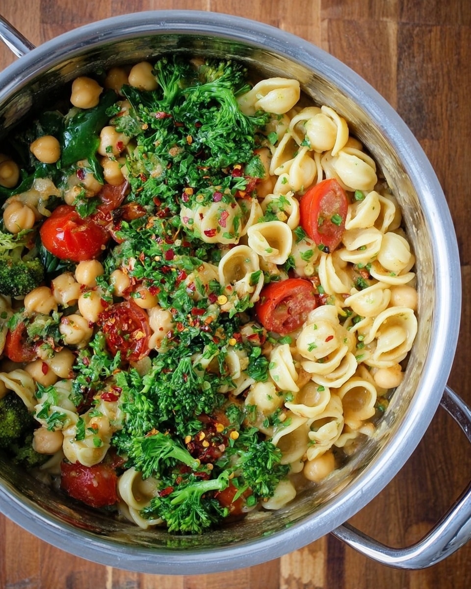 A metal cooking pot filled with a pasta dish consisting of three layers: the base layer has small round chickpeas with bright green broccoli and spinach leaves, the middle layer shows light yellow orecchiette pasta mixed evenly, and the top layer is sprinkled with chopped parsley, halved red cherry tomatoes, and small bits of red chili flakes. The pot is on a wooden surface, and the mix looks fresh and colorful with a slightly shiny texture from a light sauce. Photo taken with an iphone --ar 4:5 --v 7