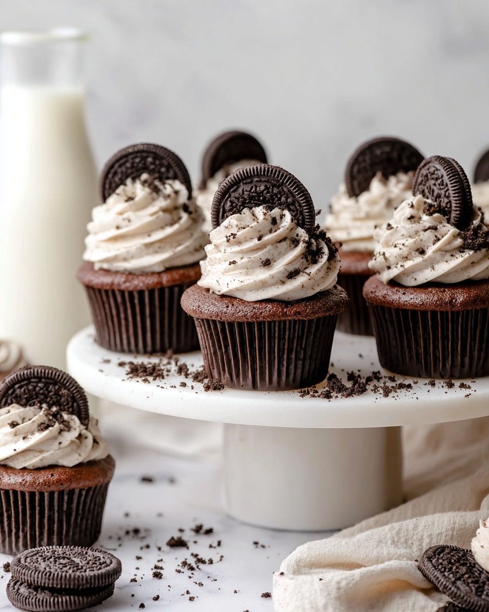 The image shows several chocolate cupcakes with a dark brown base wrapped in ridged dark brown liners. Each cupcake has two thick swirls of light grayish cream frosting speckled with tiny dark cookie bits, layered on top. A whole dark brown Oreo cookie is placed upright in the frosting on each cupcake's center, and a small amount of crushed cookie crumbs is sprinkled on the frosting surface around it. The cupcakes sit closely on a white marble board, which rests on a beige table with scattered cookie crumbs and a broken Oreo cookie in front. The background is a soft off-white color. photo taken with an iphone --ar 4:5 --v 7