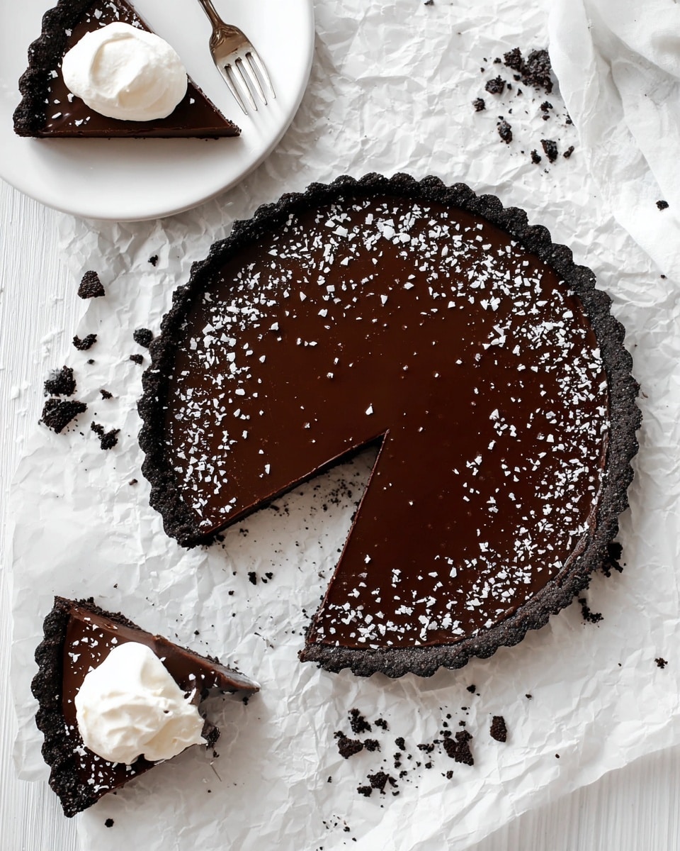 A dark chocolate tart with a shiny smooth top layer sprinkled with white flakes, showing a deep brown color, sits on crumpled white paper over a white marbled surface. The tart has a thick black crumb crust with a rough texture that forms a border around the edge. One slice is cut and placed on white paper with a dollop of white whipped cream on top, and another slice lies on a white plate in the upper left corner with a silver fork resting beside it. Crumbs are scattered around the tart and slices, creating a casual, inviting look. Photo taken with an iphone --ar 4:5 --v 7
