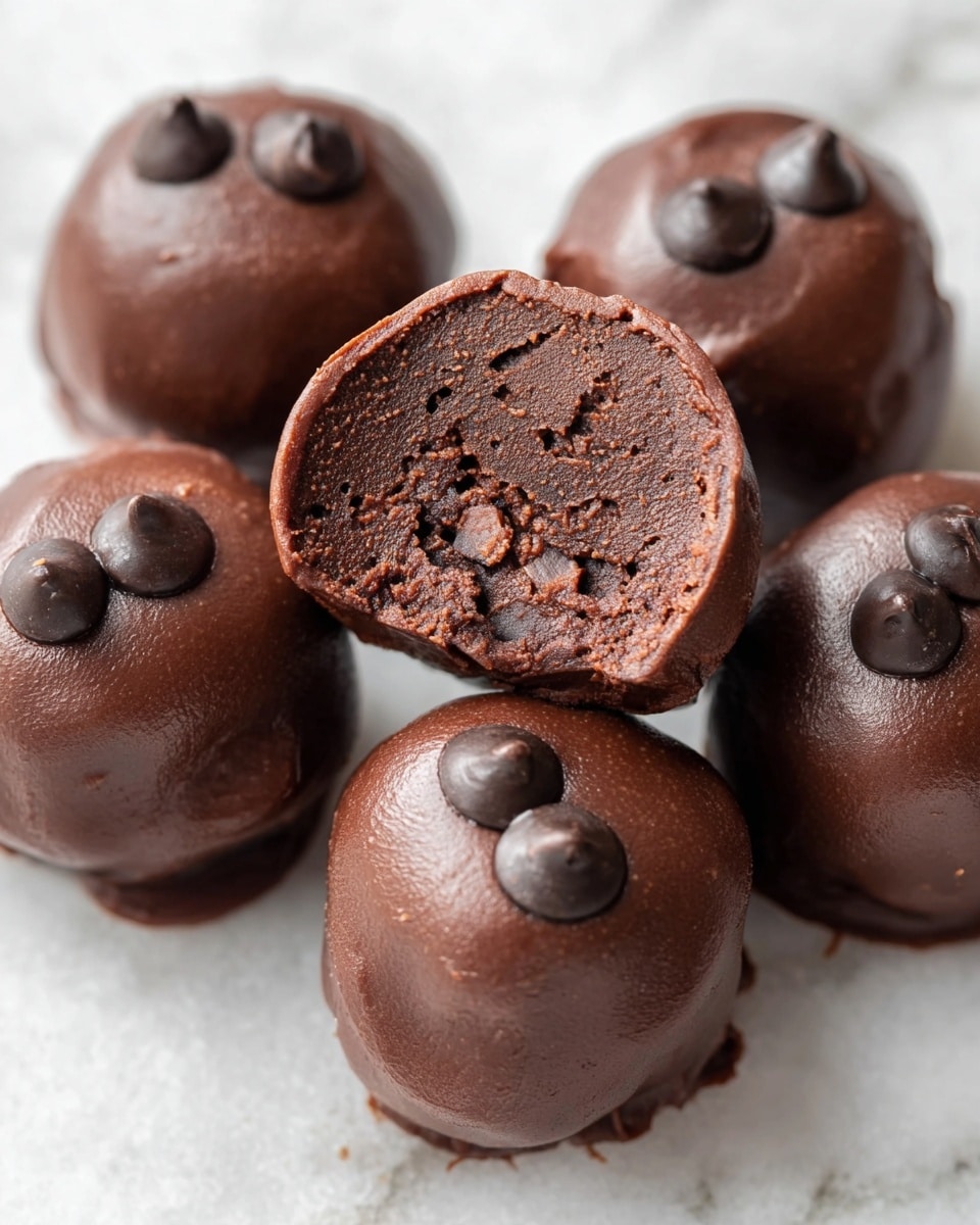 Five round chocolate truffles are placed on a white marbled surface, with one truffle in the center cut in half showing dense, rich, dark chocolate inside mixed with small pieces of chocolate chips scattered throughout. The outer layer of each truffle is smooth and shiny dark brown chocolate, with three larger, glossy chocolate chips embedded on top of each whole truffle. The texture looks soft and slightly sticky, highlighting the thick chocolate coating and filling. photo taken with an iphone --ar 4:5 --v 7