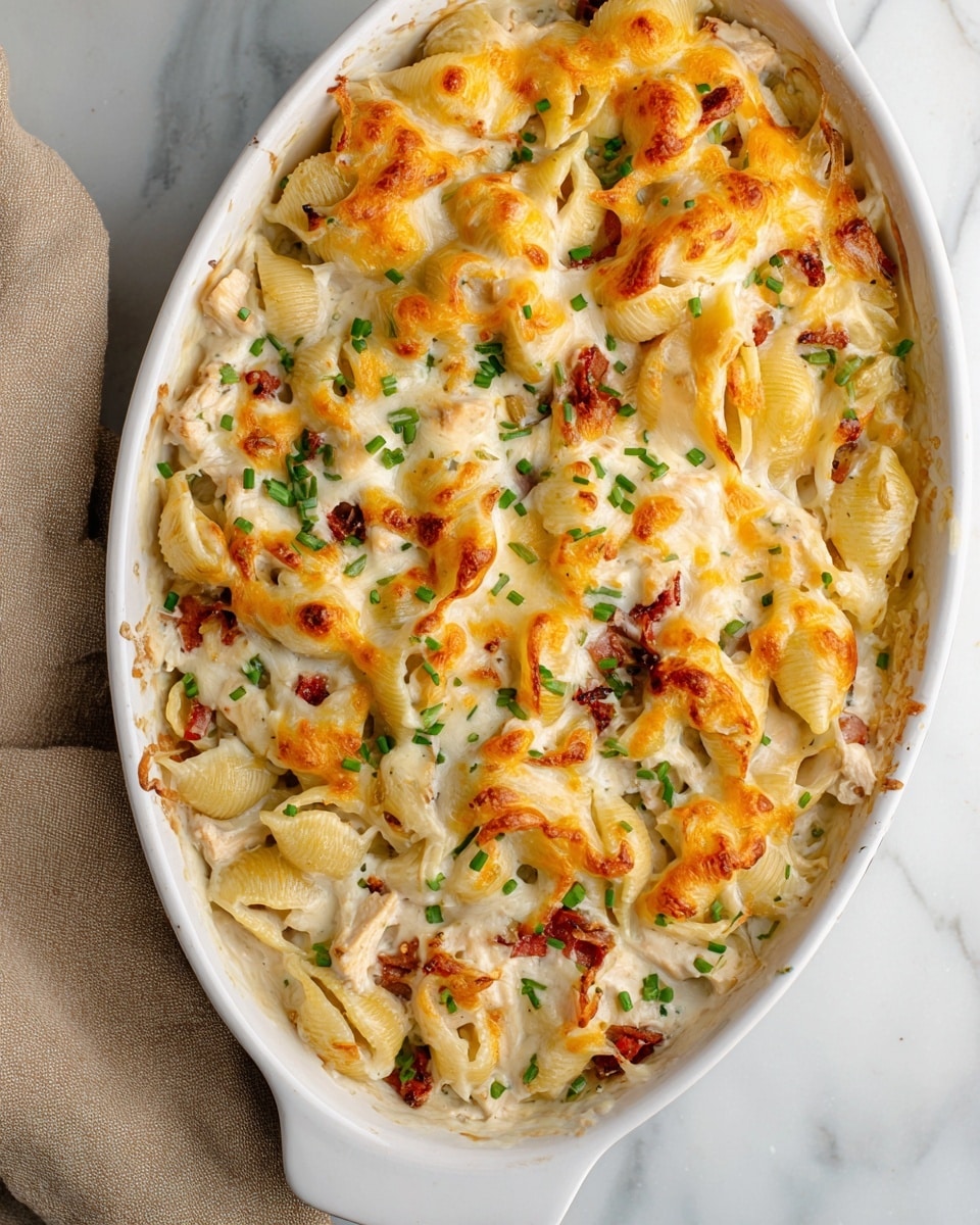 This image shows a white oval baking dish filled with a baked cheesy pasta casserole. The dish has three main layers: at the bottom, there are shell pasta pieces mixed with creamy white sauce and small bits of red, likely sun-dried tomatoes or bacon. The middle layer has chunks of white chicken meat evenly spread through the pasta. On top, melted cheese covers the entire dish, with parts of it browned and bubbly, showing a mix of white and orange-yellow cheese. Small green chopped chives are sprinkled lightly on the melted cheese, adding a fresh pop of color. The dish sits on a white marbled surface with a beige towel to the side. photo taken with an iphone --ar 4:5 --v 7