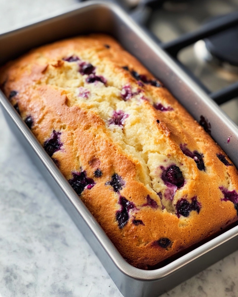 A golden brown blueberry loaf cake with a cracked top, showing soft, fluffy texture inside. The cake has purple blueberry spots spread throughout the surface and inside layers. It sits inside a rectangular silver metal baking pan placed on a stove. The cake's top has a slightly shiny and uneven crust with some cracks revealing the moist crumb beneath. The background is a white marbled texture. photo taken with an iphone --ar 4:5 --v 7