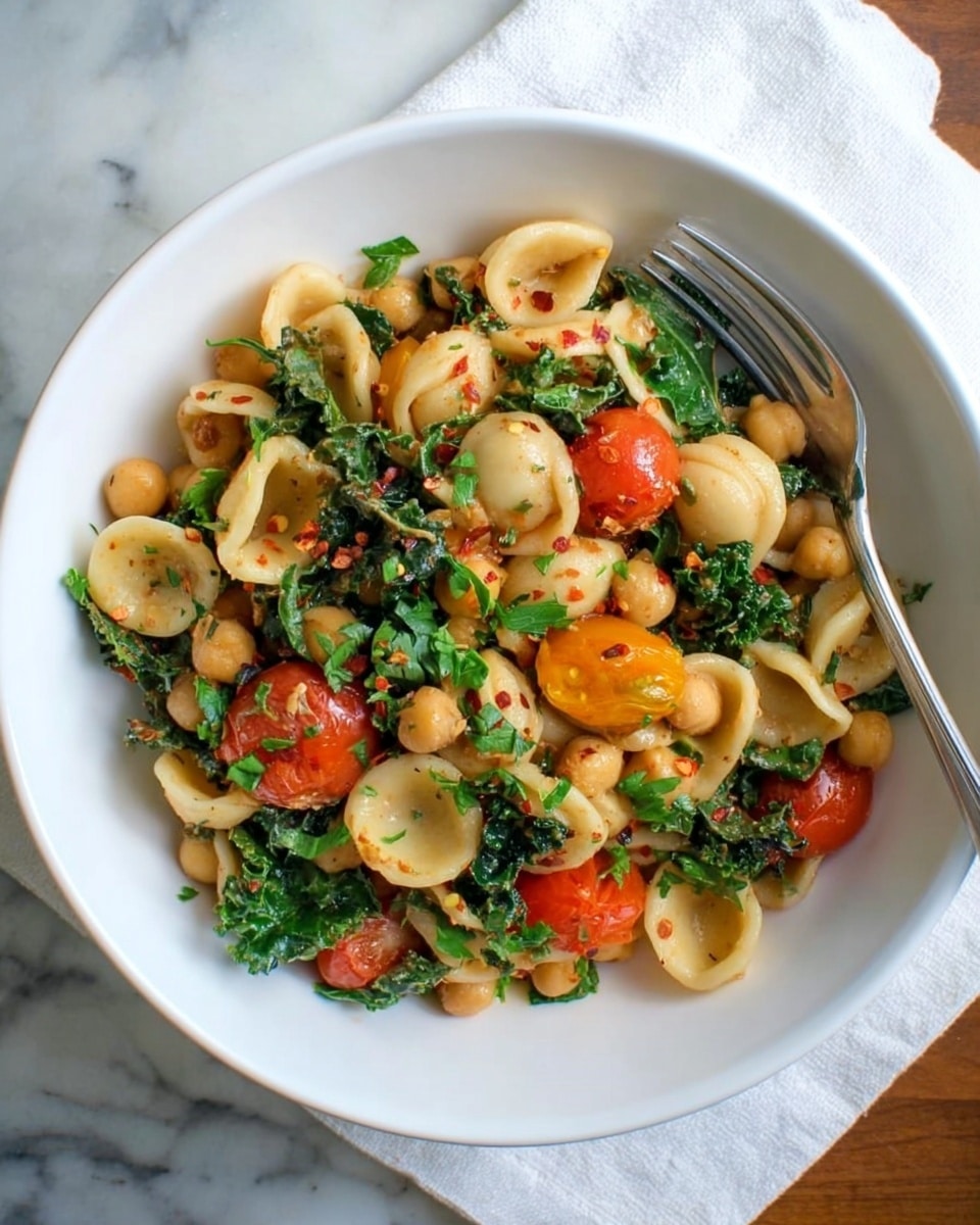A white bowl filled with orecchiette pasta mixed with chickpeas, bright green leafy kale, and halved cherry tomatoes in red and orange tones. The pasta and chickpeas are light beige, coated in a glossy sauce with small red chili flakes scattered throughout. Fresh green parsley leaves are sprinkled on top, adding brightness. A silver fork rests on the right edge of the bowl. The bowl is on a white marbled surface with a white cloth underneath. photo taken with an iphone --ar 4:5 --v 7