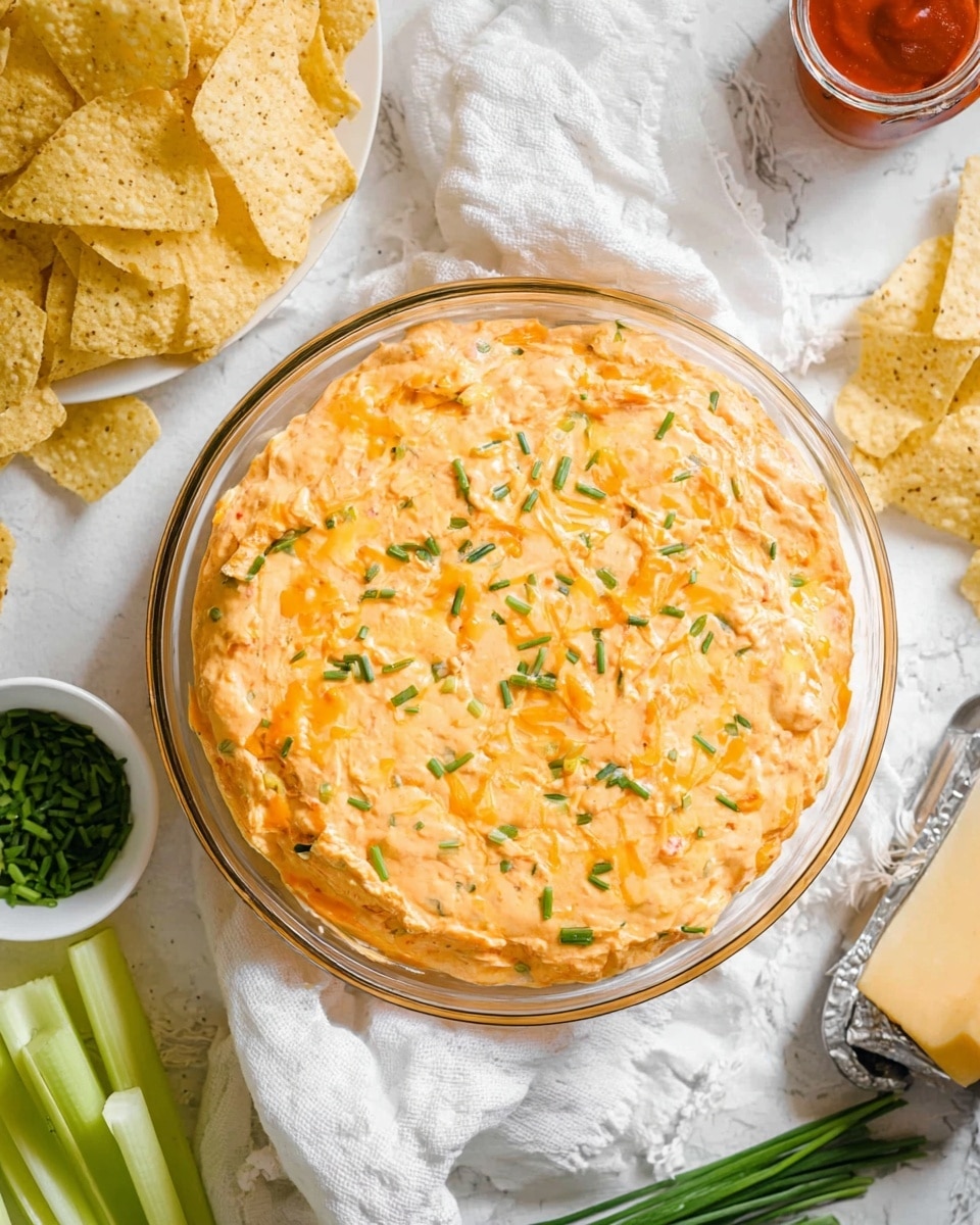 A clear glass round dish filled with a thick, creamy orange dip that looks soft and slightly chunky, topped with small green chive pieces scattered evenly over the surface; the dip fills the dish completely and shows slight browning near the edges. Around the dish is a white cloth, some celery sticks in light green are placed next to the dish, and a pile of lightly salted tortilla chips with a pale yellow color can be seen on the left side. Nearby, there is a small heap of finely chopped chives on a white dish, a shredder with a light yellow block of cheese resting on it on the right, and a small container with a red sauce visible to the left. The whole scene is set on a white marbled textured background. photo taken with an iphone --ar 4:5 --v 7
