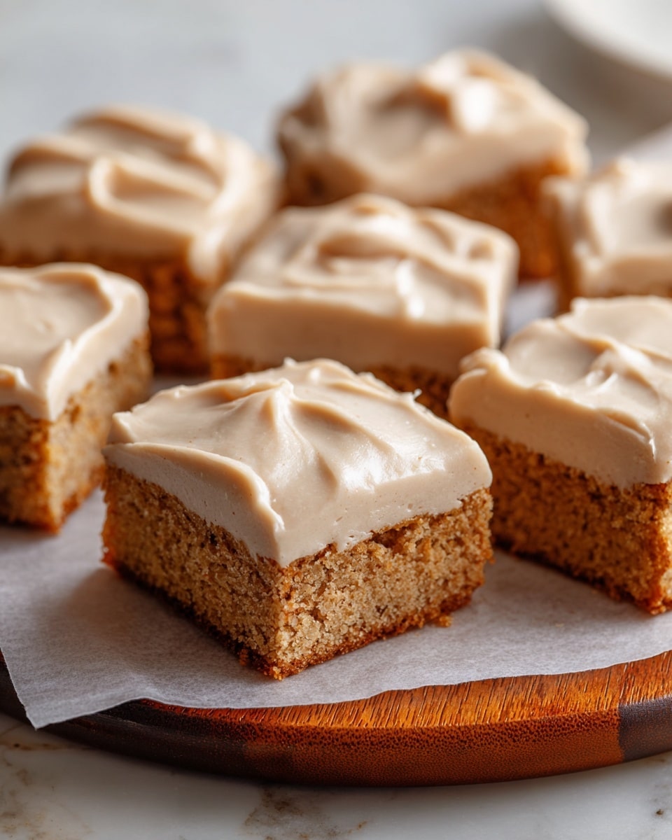 The image shows several square pieces of cake with a light brown, moist-looking crumb layer topped with a thick, creamy layer of smooth, beige frosting. Each cake square has one layer of crumb with a slightly golden crust on the bottom, and the frosting covers the top fully with soft swirls and slight peaks. The pieces are arranged closely on white parchment paper, which rests on a round wooden board, all set against a white marbled textured surface in the background. photo taken with an iphone --ar 4:5 --v 7