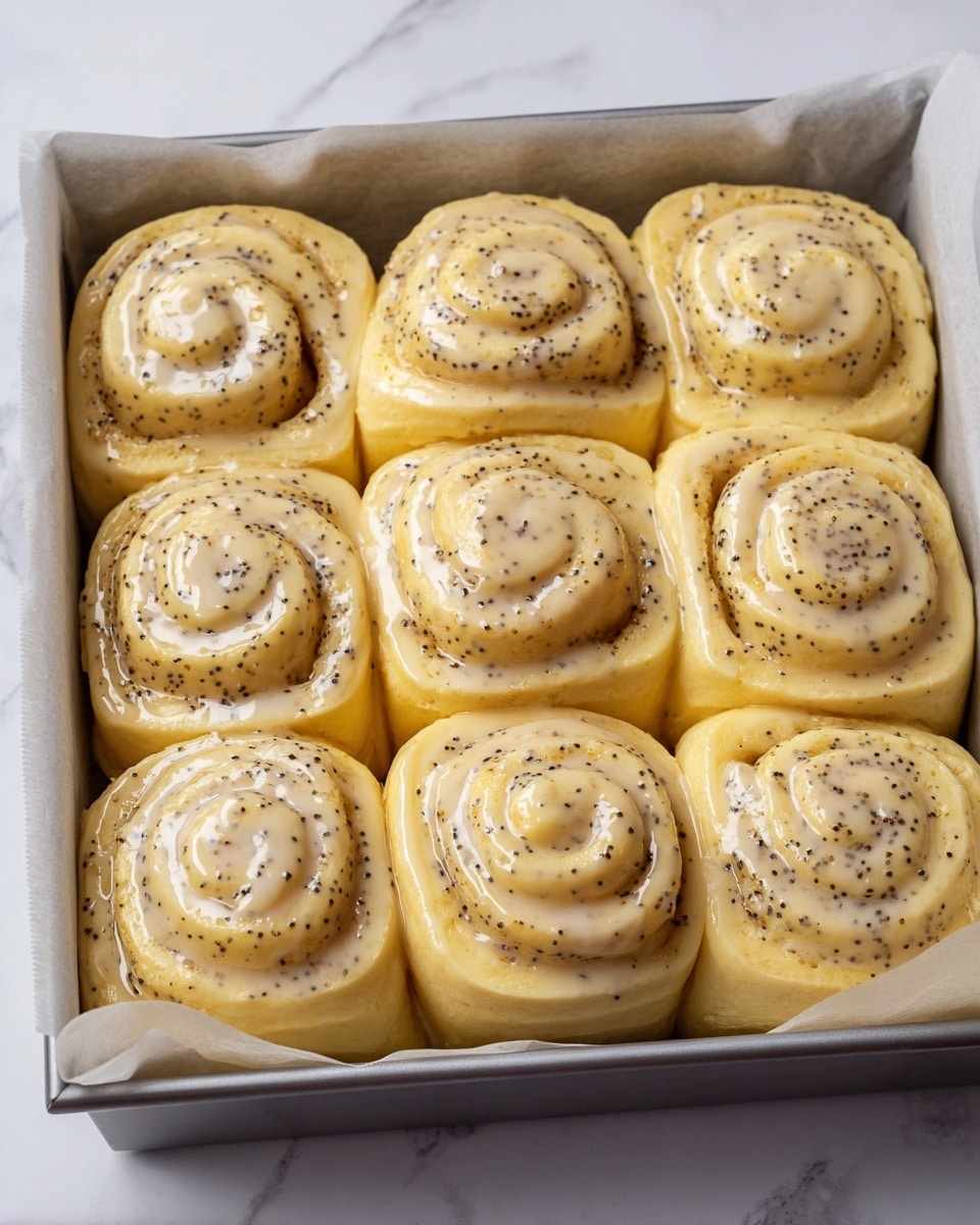 A metal baking pan lined with parchment paper holds twelve swirled rolls arranged closely together in three rows of four, each roll showing two visible layers of soft, pale yellow dough speckled with small black poppy seeds. The top layer of each roll glistens with a shiny, light glaze, and the swirls create a smooth spiral pattern. The pan rests on a white marbled surface, giving a clean and bright backdrop. photo taken with an iphone --ar 4:5 --v 7