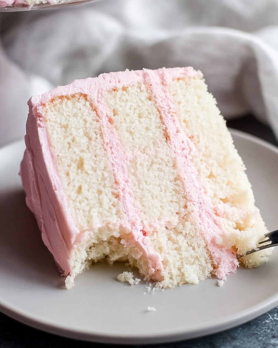 A slice of cake with three thick layers of soft white sponge separated by two layers of smooth, light pink frosting, sitting on a white plate with a smooth, matte finish. The cake has a bite taken out from the middle side, showing the moist texture inside. The surface beneath the plate is a white marbled texture with subtle gray veins, and a light-colored fabric is softly blurred in the background. Photo taken with an iphone --ar 4:5 --v 7