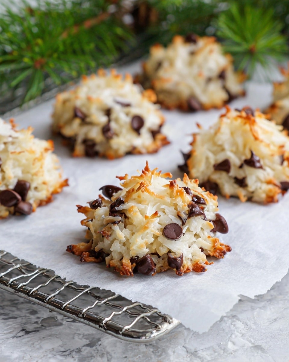 The image shows several coconut macaroons with chocolate chips on a white parchment paper that rests on a silver cooling rack. Each macaroon has one main layer made of shredded white coconut mixed with dark brown chocolate chips, giving a rough texture with some golden-brown toasted edges. The macaroons look slightly irregular in shape with small pieces of coconut sticking out. The background includes some blurred green pine branches on top of a white marbled textured surface. photo taken with an iphone --ar 4:5 --v 7