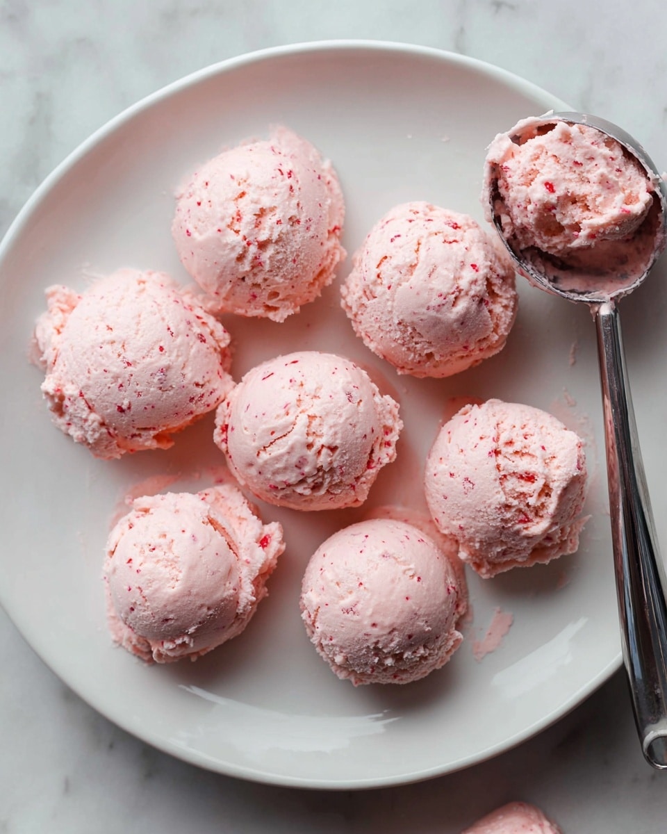 Seven scoops of light pink, slightly textured creamy mixture with small red specks are evenly spaced on a large white plate. The scoops have a soft, whipped look with rough, uneven surfaces. A metal ice cream scoop, partly filled with the same pink mixture, rests on the plate's edge on the right side. The plate is placed on a white marbled surface. photo taken with an iphone --ar 4:5 --v 7