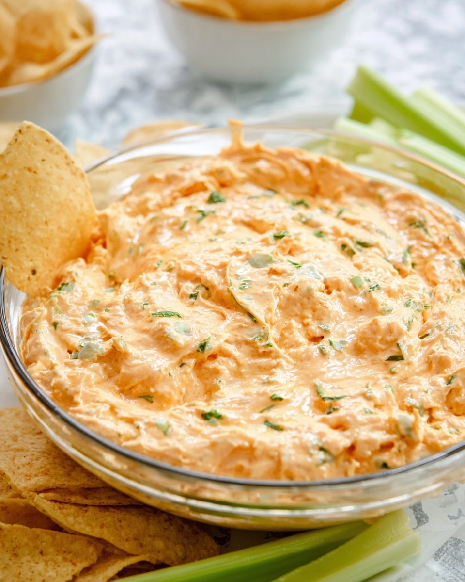 A clear glass bowl filled with a creamy, textured mixture that is light orange in color, with small green herb pieces sprinkled on top. The bowl contains several tortilla chips partially dipped into the mixture on the left side and a few celery sticks dipped on the right side. The background is a white marbled texture with some blurred round white bowls filled with dip and tortilla chips. The creamy layer inside the bowl shows slight peaks and unevenness, giving it a soft, inviting look. photo taken with an iphone --ar 4:5 --v 7