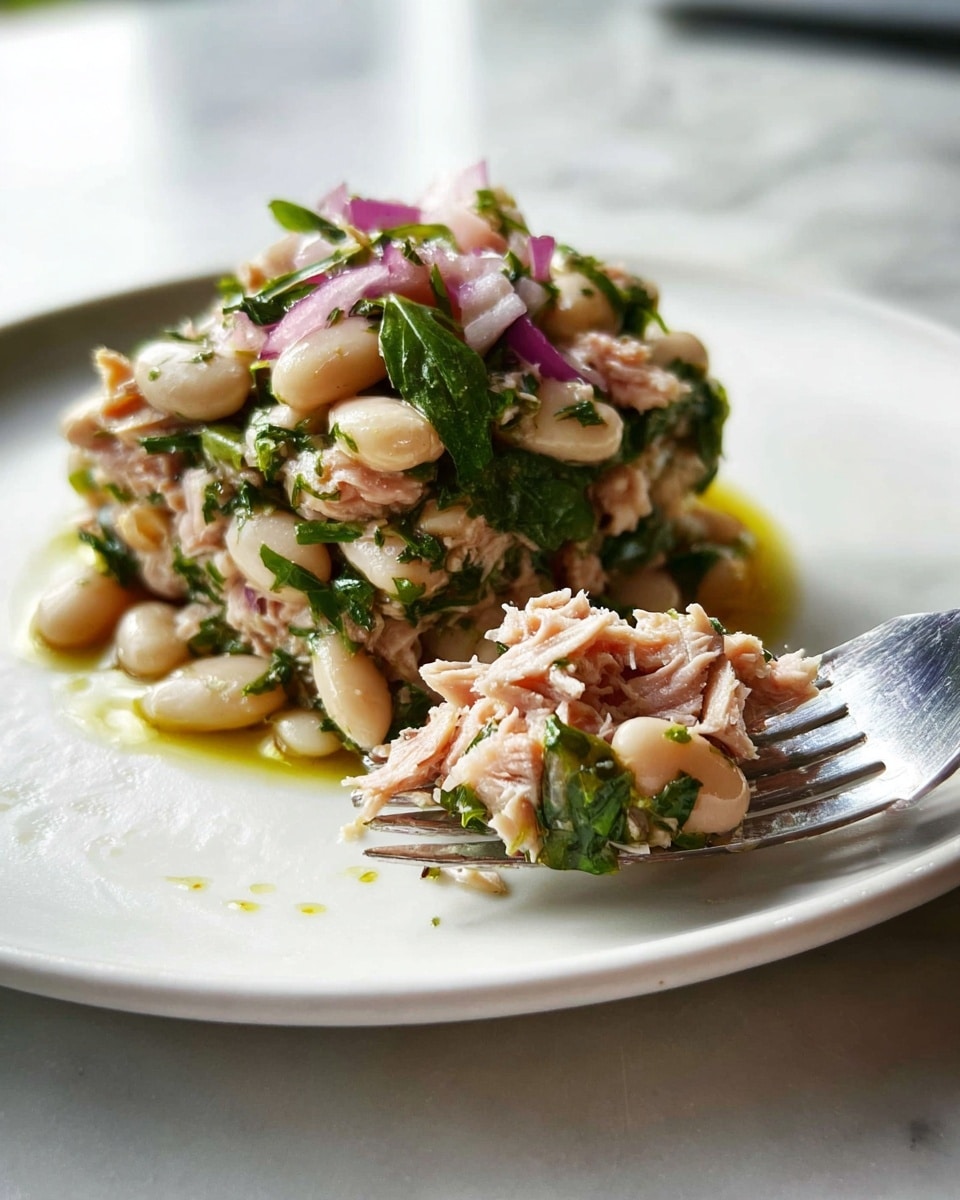 A close-up of a white plate with a pile of a layered dish made of large white beans mixed with green herbs and small pieces of red onion forming the bottom layer, topped by shredded pale pink tuna mixed with more chopped green herbs and red onions as the upper layer, all slightly glossy with olive oil, with a silver fork holding a small mix of these layers at the front right side, set on a white marbled surface, photo taken with an iphone --ar 4:5 --v 7