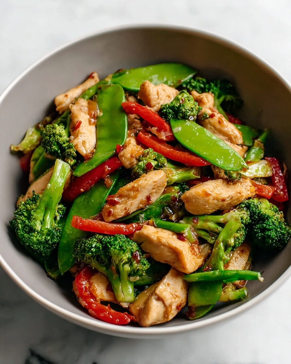 The dish is a colorful stir-fry served in a white bowl, filled with layers of light brown, tender chicken pieces that have a slightly glossy texture from the sauce. Mixed in are bright green broccoli florets with a bumpy texture, vibrant green sugar snap peas that are smooth and curved, and thin strips of red bell peppers adding a pop of red color. The layers are mixed and evenly distributed, giving a fresh and cooked look with small bits of onion peeking through. The whole bowl rests on a white marbled surface, enhancing the bright colors of the vegetables and chicken. photo taken with an iphone --ar 4:5 --v 7