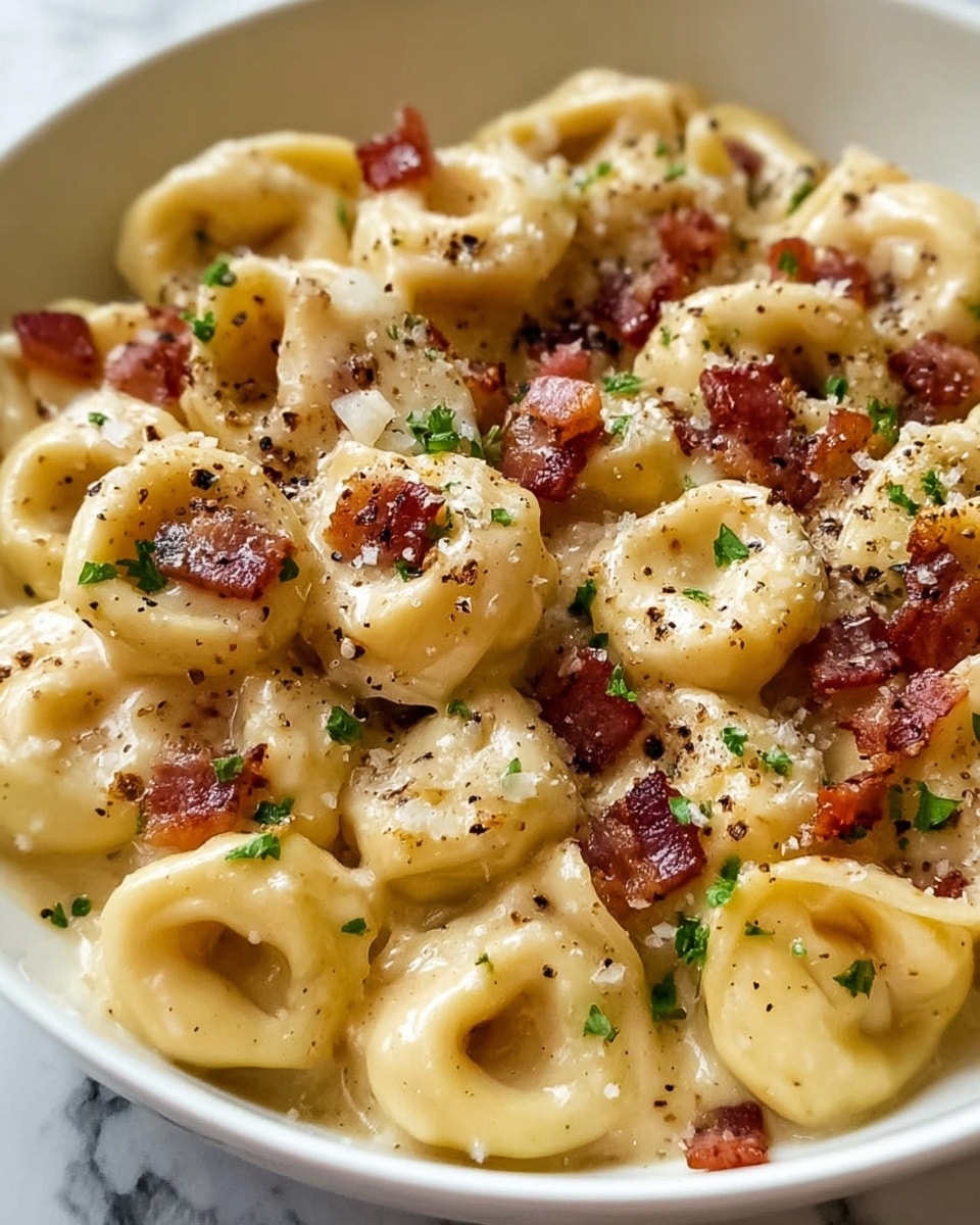 A close-up view of creamy tortellini pasta coated in a smooth, light yellow cheese sauce, filling most of a white bowl. The tortellini are round and plump, each shaped like a small ring with soft folds visible. Scattered on top are small, irregular pieces of crispy-looking reddish-brown bacon. Sprinkled over the pasta and bacon are tiny green parsley flakes and a light dusting of grated parmesan cheese, creating a slightly grainy texture. There are also small black pepper specks spread evenly on the surface, enhancing the look of seasoning. The background shows a white marbled texture. photo taken with an iphone --ar 4:5 --v 7